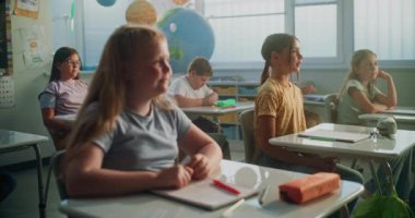 İlkokul çocukları masada oturuyor, öğretmenden ders dinliyor, doğru cevap vermek için ellerini kaldırıyor. Modern Sınıfta Coğrafya veya Bilim Öğrenen Genç Erkekler ve Kızlar Grubu.