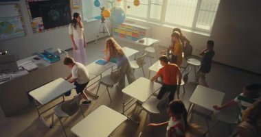 Bayan Öğretmen Okul Başında Kızları ve Oğlanları selamlıyor. İlkokul öğrencileri sınıfa giriyor, masada oturuyor ve okul malzemeleriyle sırt çantalarını açıyor. Yüksek Açı.