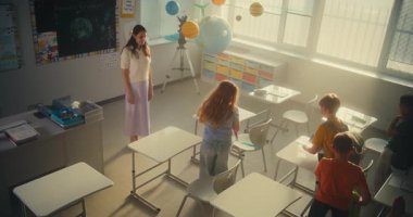 Okul Gününün Başında Kız ve Oğlanları Selamlayan Kadın Öğretmen. İlkokul öğrencileri sınıfa giriyor, masada oturuyor ve sırt çantalarını boşaltıyorlar. Ağır çekim. Yüksek Açı.