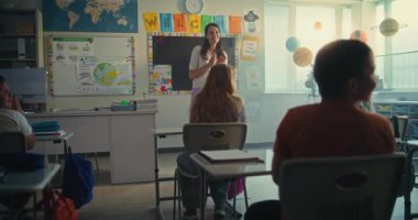 Bayan öğretmen, ilkokul çocuklarıyla dolu sınıfın önünde yeni öğrenciyi tanıtıyor. Masada oturan, el çırpan, dersten önce yeni sınıf arkadaşını selamlayan bir grup farklı çocuk..