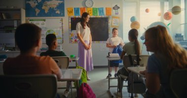 Bayan öğretmen, ilkokul çocuklarıyla dolu sınıfın önünde yeni öğrenciyi tanıtıyor. Masada oturan, el çırpan, dersten önce yeni sınıf arkadaşını selamlayan bir grup farklı çocuk..