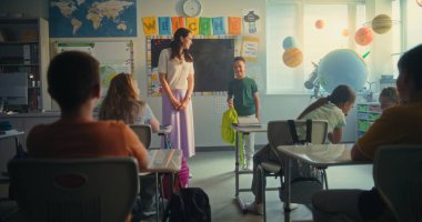 Bayan öğretmen, ilkokul çocuklarıyla dolu sınıfın önünde yeni öğrenciyi tanıtıyor. Masada oturan, el çırpan, dersten önce yeni sınıf arkadaşını selamlayan bir grup farklı çocuk..