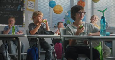 İlkokul çocukları masada oturuyor, beslenme çantalarından sandviç yiyor, teneffüste birbirleriyle konuşuyorlar. Modern Renkli Sınıflar Dersinden Önce Mutlu Çeşitli Çocuklar Öğle Yemeği Zamanları.