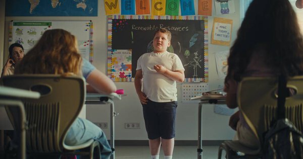 Застенчивый первоклассник, демонстрирующий знания об экологии перед классом, полным детей в современном классе. Женщина-преподаватель поддерживает молодых студентов во время презентации. STEM Образование. Slow Motion.