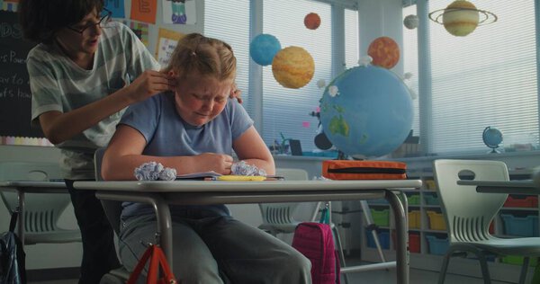 Depressed Primary School Girl Sitting Alone at Desk in Classroom While Aggressive Classmates Abusing Her, Throwing Papers and Laughing. School Bullying, Peer Harassment and Toxic School Environment.