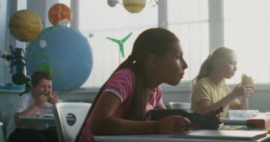 Masada oturan, öğle yemeği kutusundan leziz kurabiyeler yiyen ve molada sınıf arkadaşlarıyla konuşan genç bir kız. İlkokul öğrencileri Modern Parlak Sınıfta Ders Öncesi Öğle Yemeği Yiyor