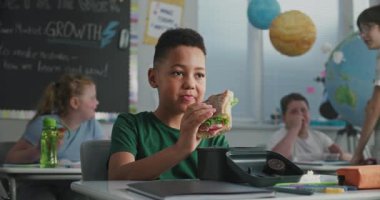 Afrika kökenli Amerikalı ilkokul çocuğu, yemek kutusundan sandviç yiyor, teneffüs sırasında sınıf arkadaşıyla konuşuyor. Çok Irklı Sınıflar Dersinden Önce Mutlu Çeşitli Çocuklar Yemek Yiyor.