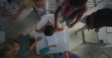 Depresyondaki ilkokul çocuğu masada oturmuş, agresif sınıf arkadaşları onu taciz ederken kameraya bakıyor, kağıtları fırlatıyor ve gülüyor. Okul zorbalığı ve Toksik Okul Ortamı. Üst Görünüm Döndürülüyor.