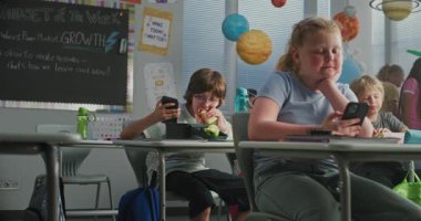 İlkokul çocuklarının sandviç yemesi, akıllı telefonlar kullanması ve tatilde birbirleriyle konuşması. Modern Renkli Sınıflar 'da, Masalarda oturan, Dersten Önce Öğle Yemeği Yiyen Mutlu Çocuklar.