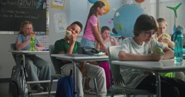 Afro-Amerikan İlköğretim Okulu öğrencisi masada oturuyor, Beslenme Kutusundan Ev Yapımı Sandviç yiyor, Sınıf arkadaşıyla konuşuyor. Modern Sınıfta Ders Öncesi Öğle Yemeği Yiyen Genç Erkekler ve Kızlar