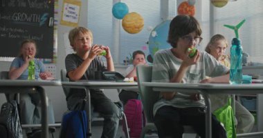 İlkokul çocukları masada oturuyor, beslenme çantalarından sandviç yiyor, teneffüste birbirleriyle konuşuyorlar. Modern Renkli Sınıflar Dersinden Önce Mutlu Çeşitli Çocuklar Öğle Yemeği Zamanları.