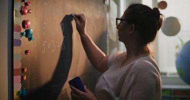 İlkokul öğrencilerine ders sırasında matematik problemlerini çözmeyi öğreterek, Chalkboard 'a kadın öğretmen yazma ve çarpma tablosu. Renkli Sınıfta Matematik Sınıfı. Kapat..