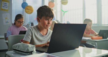 Bilgisayar Bilimi Dersi: İlkokul çocuğu masada oturuyor, sınıfta dizüstü bilgisayar kullanıyor. Temel Programlama Dillerini Öğrenen Erkekler ve Kızlar Grubu ve Dijital Tabletler ve Dizüstü bilgisayarlarda İnternet Güvenliği.