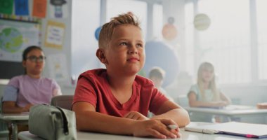 İlkokul çocuğu masada oturuyor, elini kaldırıyor ve coğrafya dersi sırasında öğretmenlere soru soruyor. Modern Renkli Sınıfta Çevre Bilimi okuyan Zeki Çeşitli Çocuklar Takımı.