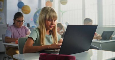 Masada oturan ilkokul öğrencisi kız, sınıfta dizüstü bilgisayar kullanıyor. Bilgisayarları ve Tablet Bilgisayarları üzerinde çalışan, Bilgisayar Bilimi, Temel Programlama ve Kodlama okuyan Zeki Çeşitli Çocuklar Takımı.