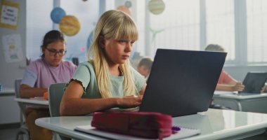 Bilgisayar Bilimi Dersi: İlkokul öğrencisi kız masamda oturuyor, sınıfta dizüstü bilgisayar kullanıyor. Temel Programlama Dillerini Öğrenen Erkekler ve Kızlar Grubu ve Dijital Tabletler ve Dizüstü bilgisayarlarda İnternet Güvenliği.