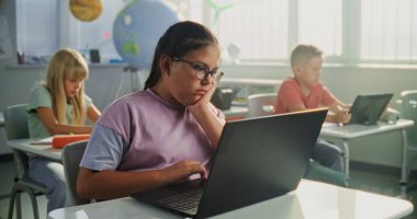 Bilgisayar Bilimi Dersi: İlkokul öğrencisi kız masamda oturuyor, sınıfta dizüstü bilgisayar kullanıyor. Temel Programlama Dillerini Öğrenen Erkekler ve Kızlar Grubu ve Dijital Tabletler ve Dizüstü bilgisayarlarda İnternet Güvenliği.