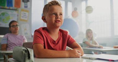 İlkokul çocuğu masada oturuyor, elini kaldırıyor ve coğrafya dersi sırasında öğretmenlere soru soruyor. Modern Renkli Sınıfta Çevre Bilimi okuyan Zeki Çeşitli Çocuklar Takımı.