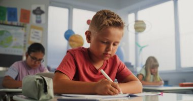 İlkokul çocuğu masada oturuyor, Okul Defteri Üzerine Ders Yazıyor. Modern Renkli Sınıfta Coğrafya veya Çevre Bilimi Üzerine Yazı Yazan Genç Erkekler ve Kızlar Grubu.