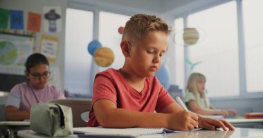 İlkokul çocuğu masada oturuyor, Okul Defteri Üzerine Ders Yazıyor. Modern Renkli Sınıfta Coğrafya veya Çevre Bilimi Üzerine Yazı Yazan Genç Erkekler ve Kızlar Grubu.