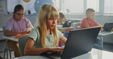 Masada oturan ilkokul öğrencisi kız, sınıfta dizüstü bilgisayar kullanıyor. Bilgisayarları ve Tablet Bilgisayarları üzerinde çalışan, Bilgisayar Bilimi, Temel Programlama ve Kodlama okuyan Zeki Çeşitli Çocuklar Takımı.