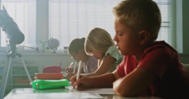 İlkokul çocukları masada oturuyor, okul sınavı yazıyor ya da not defterlerinde görevler yapıyor. Modern Sınıfta Genç Erkek ve Kız Yazarlık Testi, Çevre Bilimi ve Coğrafyası Öğrenme Grubu.