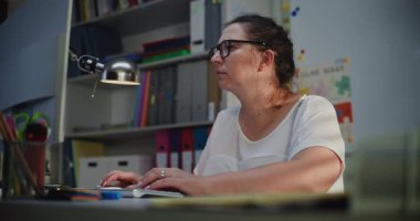 İlkokul öğretmeni Bilgisayar 'da çalışıyor, Ödevleri kontrol ediyor ve Uzaktan Öğrenme, Öğrenciler için Ders Hazırlama Planı sırasında online görevleri yapıyor. Akşam vakti Boş Sınıfta Bilgisayarda Çalışan Kadın.