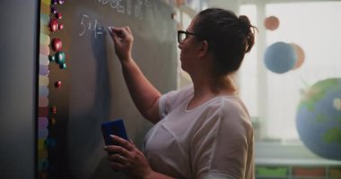 Tebeşir tahtasında yazan, öğrencilerle konuşan, matematik dersi sırasında ilkokul çocuklarına sayıları çarpma yöntemini açıklayan kadın öğretmen. Modern Renkli Sınıflar Matematik Sınıfı