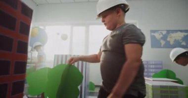 Hardhat şapkalı Zeki Çocuklar Karton Şehir Modelleri, Yollar ve Ağaçlarla Etkileşimli Şehir Planlaması Yapıyor. Bayan öğretmen, ilkokul çocukları için BTMM etkinliği hazırlıyor. El bilgisayarı.