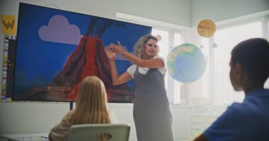 Jeoloji sınıfı: Dijital ekranı kullanarak ilkokul çocuklarına öğretmenlik yapan kadın öğretmen. Masalarda oturan, Volkan Patlaması ve Modern Sınıfta Coğrafya Öğrenen Genç Öğrenciler Grubu.