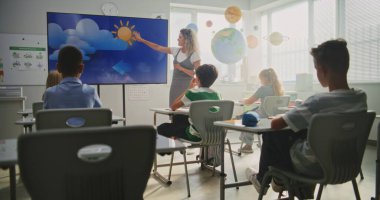 Coğrafya Sınıfı: Dijital Ekran Kullanarak İlkokul Çocuklarına Öğretmenlik Yapan Kadın Öğretmenler. Genç Erkekler ve Kızlar Masada Oturuyor, Modern Sınıfta Su Döngüleri ve Hava Durumu Faktörlerini Öğreniyor