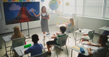 Jeoloji sınıfı: Dijital ekranı kullanarak ilkokul çocuklarına öğretmenlik yapan kadın öğretmen. Masalarda oturan, Volkan Patlaması ve Modern Sınıfta Coğrafya Öğrenen Genç Öğrenciler Grubu.