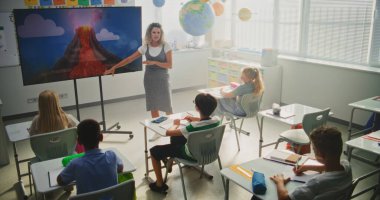 Jeoloji sınıfı: Dijital ekranı kullanarak ilkokul çocuklarına öğretmenlik yapan kadın öğretmen. Masalarda oturan, Volkan Patlaması ve Modern Sınıfta Coğrafya Öğrenen Genç Öğrenciler Grubu.