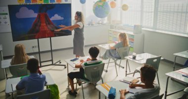 Jeoloji sınıfı: Dijital ekranı kullanarak ilkokul çocuklarına öğretmenlik yapan kadın öğretmen. Masalarda oturan, Volkan Patlaması ve Modern Sınıfta Coğrafya Öğrenen Genç Öğrenciler Grubu.