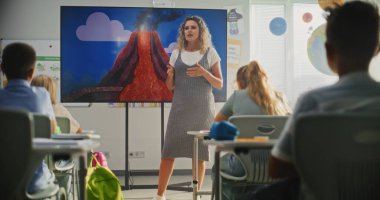 Jeoloji sınıfı: Dijital ekranı kullanarak ilkokul çocuklarına öğretmenlik yapan kadın öğretmen. Masalarda oturan, Volkan Patlaması ve Modern Sınıfta Coğrafya Öğrenen Genç Öğrenciler Grubu.