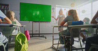 Tablet Bilgisayarlı Bayan Öğretmen İlkokul Öğrencilerini Sınıfta Eğitiyor. Büyük Dijital Ekran ve Krom Anahtar. Eğitim İçeriği için Yeşil Ekran Şablonu. Doli Vuruşu.