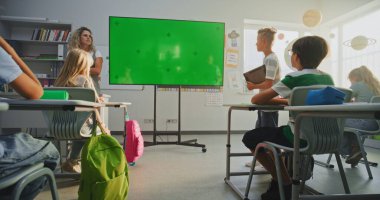 Tablet Bilgisayarlı İlkokul Çocuğu Sınıf Arkadaşlarının Önünde Sunum Yapıyor ve Öğretmen Kroma Anahtarlı Büyük Dijital Ekran Kullanıyor. Eğitim İçeriği için Yeşil Ekran Şablonu.