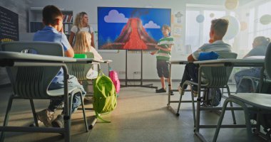 Tablet bilgisayarlı ilkokul çocuğu sınıfın önünde jeoloji bilgisi gösteriyor ve öğretmen dijital ekranı kullanıyor. Öğrenciler Sunum Dinliyor, Volkanik Patlama Sürecini Öğreniyor.