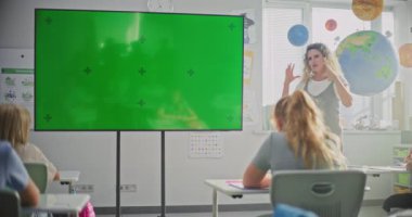 Modern Sınıflar: Chromakey Mockup ile Büyük Dijital Ekran kullanarak İlkokul Çocuklarına Öğretmenlik Yapan Kadın Sınıfı. Eğitim İçeriği veya Etkileşimli Görsel Efektler için Yeşil Ekran Yer Tutucu.