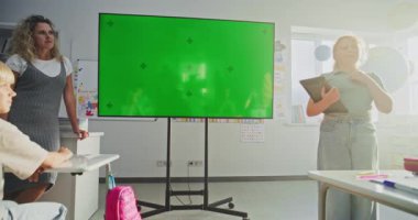 Tablet Bilgisayarlı İlkokul Kızı Sınıf Arkadaşlarının Önünde Sunum Yapıyor ve Öğretmen Kroma Anahtarlı Büyük Dijital Ekran Kullanıyor. Eğitim İçeriği için Yeşil Ekran Şablonu.