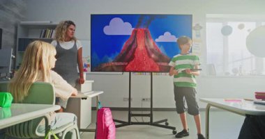 Tablet bilgisayarlı ilkokul çocuğu sınıfın önünde jeoloji bilgisi gösteriyor ve öğretmen dijital ekranı kullanıyor. Öğrenciler Sunum Dinliyor, Volkanik Patlama Sürecini Öğreniyor.