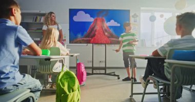 Tablet Bilgisayarlı İlkokul çocuğu ödevini sunuyor, sınıf arkadaşlarının önündeki volkanik püskürme sürecini ve dijital ekranı kullanan öğretmeni anlatıyor. Sınıfta Jeoloji okuyan Zeki Çocuklar.