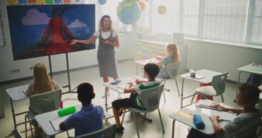 Jeoloji sınıfı: Dijital ekranı kullanarak ilkokul çocuklarına öğretmenlik yapan kadın öğretmen. Masalarda oturan, Volkan Patlaması ve Modern Sınıfta Coğrafya Öğrenen Genç Öğrenciler Grubu.