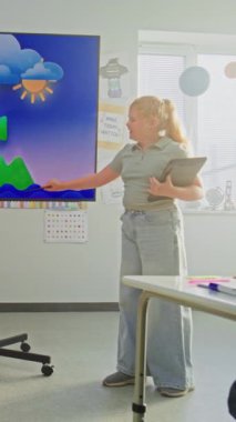 Tablet bilgisayarlı ilkokul kızı sınıfın önünde coğrafya bilgisi gösteriyor ve öğretmen dijital ekranı kullanıyor. Genç Öğrenciler Su Döngüleri ve Hava Durumu Faktörleri Öğreniyor. Dikey Çekim.