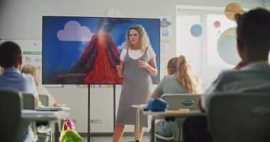 Jeoloji sınıfı: Dijital ekranı kullanarak ilkokul çocuklarına öğretmenlik yapan kadın öğretmen. Masalarda oturan, Volkan Patlaması ve Modern Sınıfta Coğrafya Öğrenen Genç Öğrenciler Grubu.