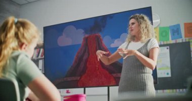 Jeoloji sınıfı: Dijital ekranı kullanarak ilkokul çocuklarına öğretmenlik yapan kadın öğretmen. Masalarda oturan, Volkan Patlaması ve Modern Sınıfta Coğrafya Öğrenen Genç Öğrenciler Grubu.