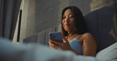 Güzel Kız Güneşli Sabah Kahvaltısını Yatakta Yiyor ve İnternetten Sohbet Ediyor. Asyalı Kadın İnternette Ceviz ve İletişim 'i seviyor. Sabah Rutini Konsepti, Yatakta Kahvaltı, Dijital Hayat. El bilgisayarı.