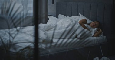 Modern Sahne, Sıcak Yatak Odası. Genç Asyalı Kadın Beyaz Yatağa Uzanıyor ve Yumuşak Battaniyenin Altında Yatıyor. Zor bir günden sonra rahatlama, sağlıklı uyku ve iyileşme, kişisel bakım kavramı. El bilgisayarı.