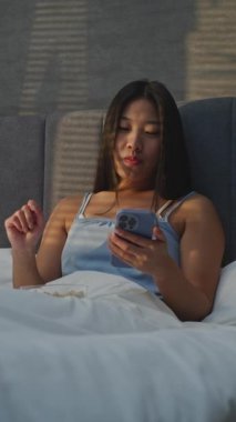 Yatakta oturan, kahvaltıda fındık yiyen ve haberciyle sohbet eden kız. Güzel Asyalı Kadın Yatak Odasında Yemek Yemekten Hoşlanıyor, İnternette Sohbet Ediyor. Sabah rutini, Yatakta Kahvaltı, Dijital Hayat. Dikey Çekim.