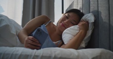 Güzel Asyalı Kız Yatak Odasında Boş Zaman Geçiriyor. Genç Kadınlar Yatakta Uzanıyor Haber Kaynağı Arasında Akıllı Telefon ve Mutlu Gülümsemeler Üzerine Yazılar Yazıyor. Kavram Sabah Rutini, Sosyal Yaşam, Dijital Yaşam Tarzı. El bilgisayarı.